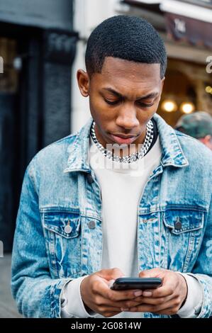 Front view of black positive young man in jeans outfit messaging on mobile phone while walking in city Stock Photo