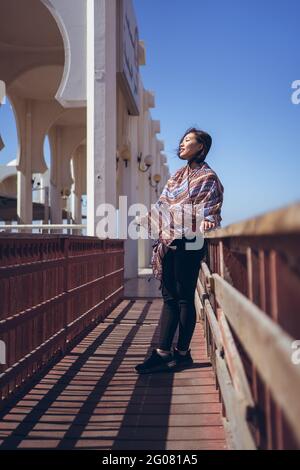 Positive young Asian female in colorful traditional headscarf smiling and looking away while standing near beautiful white building of Al Rahma Mosque Stock Photo