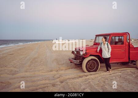 Asian Woman leaning against a red car on the beach at Turtle Beach, South, Oman Stock Photo