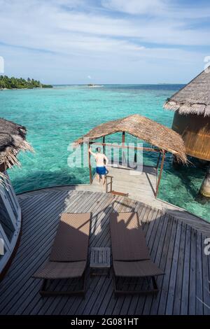 From above back view of unrecognizable male in casual swim shorts standing on wooden pier relaxing in Maldives Stock Photo