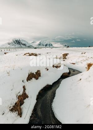 Narrow creek with cold water flowing under snow against mountains and overcast sky on Lofoten Islands, Norway Stock Photo