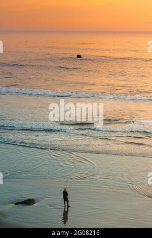 From above amazing scenery with lonely unrecognizable fisher on wet sand shore washing by foam ocean waves under colorful sky during sunset Stock Photo