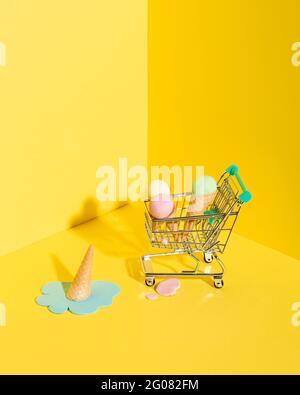 From above composition of miniature shopping trolley with assorted multicolored ice cream cones placed near fallen melting blue ice cream on yellow ba Stock Photo