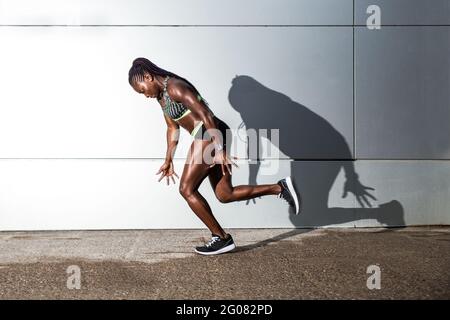 Muscular African American female in sportswear looking at camera and jumping while working out on city street near modern building wall Stock Photo