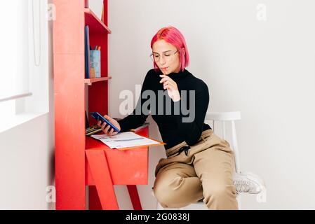 Young creative female designer with pink hair in casual outfit and glasses using smartphone and drawing sketch while working at desk at home Stock Photo