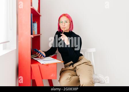 Young creative female designer with pink hair in casual outfit and glasses looking at camera using smartphone and drawing sketch while working at desk Stock Photo