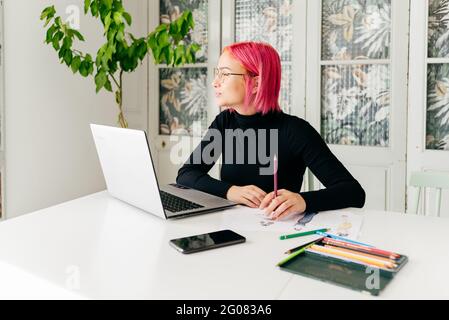 Young creative female fashion designer in casual outfit and glasses looking away pensively while sitting at table with gadgets and drawing sketch with Stock Photo