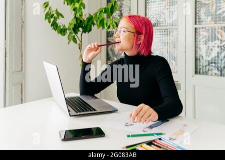 Thoughtful female freelance designer in casual outfit and glasses working at table with laptop and drawing fashion sketches with pencils Stock Photo