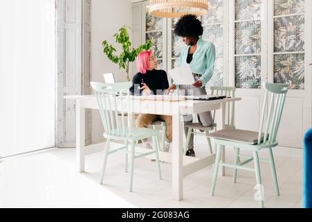 Young multiracial female fashion designers discussing sketches for new collection while working together at table in light room Stock Photo