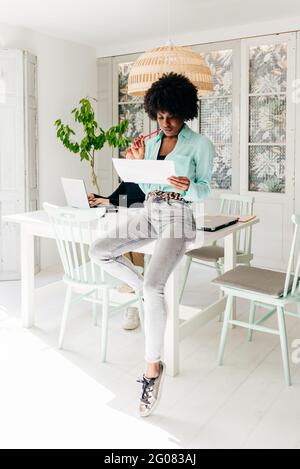 Modern successful African American female freelancer in stylish outfit with afro hair sitting at table and reading document at home Stock Photo