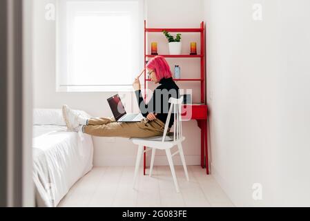 Side view of focused young female freelancer in casual clothes sitting on chair and using laptop while working on project in light modern apartment Stock Photo