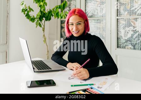 Young creative female fashion designer in casual outfit and glasses looking at camera smiling while sitting at table with gadgets and drawing sketch w Stock Photo