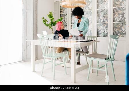 Young multiracial female fashion designers discussing sketches for new collection while working together at table in light room Stock Photo