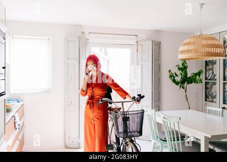 Young creative female designer in trendy outfit and eyeglasses talking on smartphone while standing with bicycle in modern light apartment Stock Photo