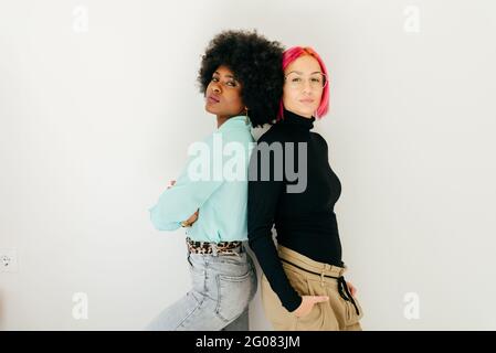 Young pink haired Woman and African American girlfriend in stylish outfit standing together back to back on white background Stock Photo