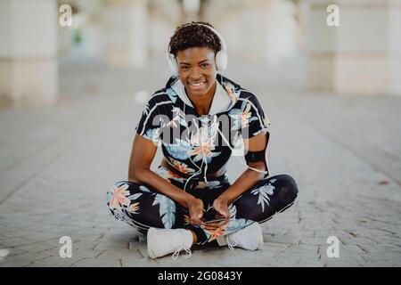 Young fitness African American Woman resting and stretching legs while listening to music after running on city street Stock Photo