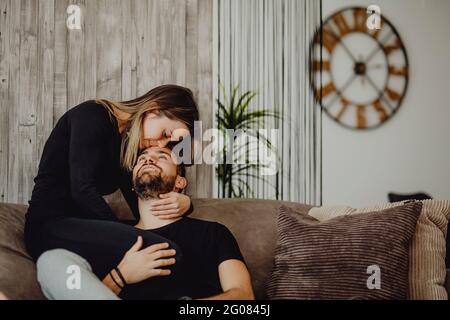 Smiling female embracing and kissing cheerful man in forehead while sitting on comfortable couch at home together Stock Photo