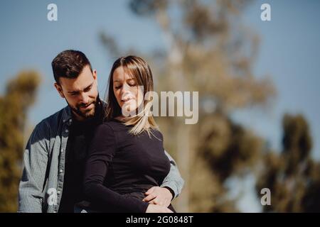 Bearded man embracing Woman while spending time on green lawn on street together Stock Photo