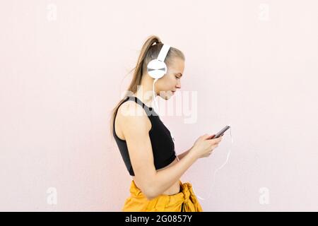 Young caucasian Woman wearing headphones and sport outfit, listening to music on the phone, isolated on bright background Stock Photo
