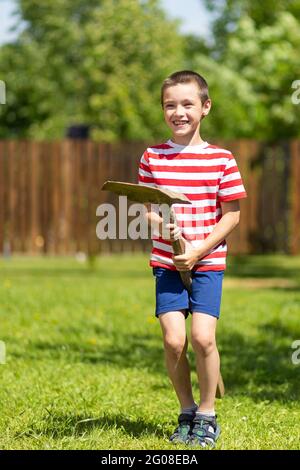 A little cheerful boy stands and sits on a shovel like a horse and jumps merrily, ready to work in the garden of a country house. Stock Photo