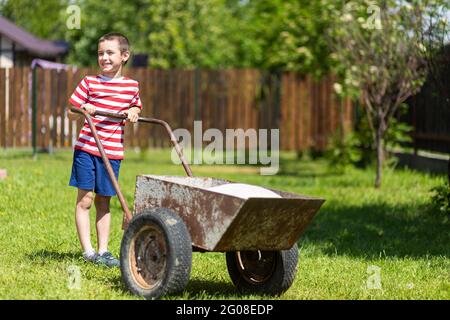 Young smile boy pushes a wheelbarrow around a yard.Boy helper in t-short and shorts having fun pushing barrow and gardening at countryside Stock Photo