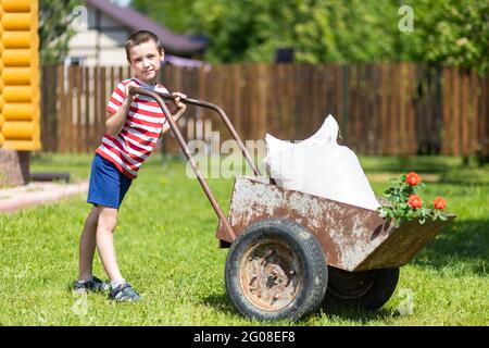 A young smiling boy is pushing a wheelbarrow across the yard. Assistant boy in t-shirt and shorts having fun pushing wheelbarrow with sack and seedlin Stock Photo