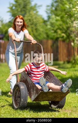 Happy cheerful boy having fun in a wheelbarrow pushing mom in home garden on a warm sunny day. Active outdoor games for children in summer. Stock Photo