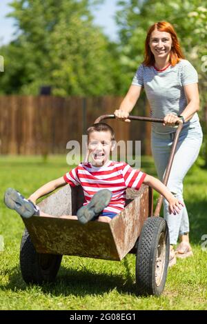 Happy cheerful boy having fun in a wheelbarrow pushing mom in home garden on a warm sunny day. Active outdoor games for children in summer. Stock Photo