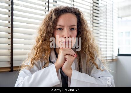 Serious female doctor carefully listening and keeping hands under chin while making medical conference video call from hospital, telehealth concept Stock Photo