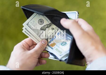 Purse with dollars in their hands. A male businessman opens a black wallet and pulls out a hundred-dollar bill on a green background. Employee salary. Stock Photo
