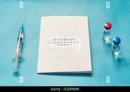 A document for recording vaccinations made in Russia, a syringe and a bottle of vaccine on a blue background. Translated from Russian: Certificate of Stock Photo