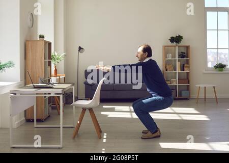 Mature businessman doing squat exercise front of laptop computer in office Stock Photo