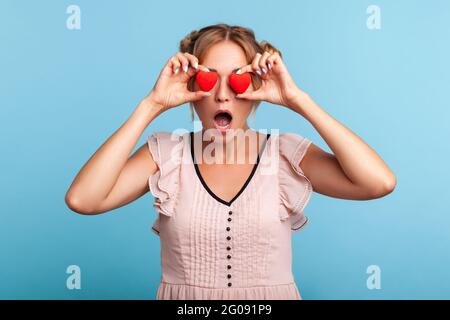 Surprised blonde woman with two funny hair buns in summer dress covering eyes with red hearts, hiding behind symbol of love, sees something astonished Stock Photo