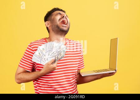 Extremely happy excited man in striped t-shirt screaming and laughing holding laptop and lot of dollar cash, enjoying jackpot winning. Indoor studio s Stock Photo