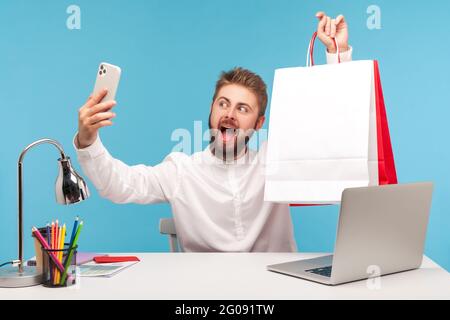 Positive bearded man blogger making selfie or recording video, posing at smartphone camera with shopping bags, bragging with purchases. Indoor studio Stock Photo