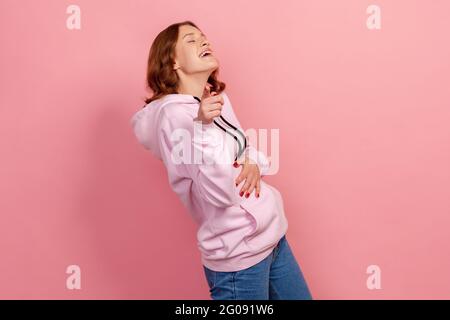Portrait of positive excited curly haired teenage girl in hoodie laughing out loud holding belly and pointing finger on you, mockery. Indoor studio sh Stock Photo