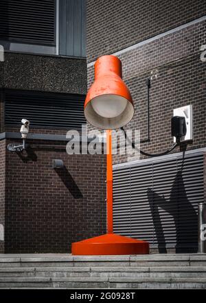 Giant enormous bright red desk lamp light with big bulb plugged into external wall of building at top of steps. Towering above brick wall on summer Stock Photo