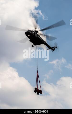 Silhouetted flying helicopter using long sling to carry small vehicle with cloudy blue sky as background. Stock Photo