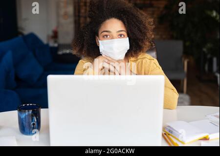 African young woman sits at the desk wearing protective mask against coronavirus and flu using laptop computer in the home office, working remotely with face covered Stock Photo