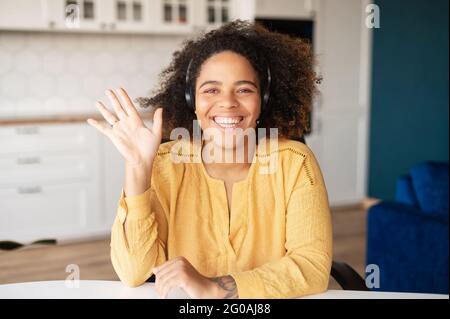 Video meeting with call center representative. Headshot of charming smiling African woman in headset, she is looking and waving at the camera. Video call to female colleague or friend, webcam view Stock Photo
