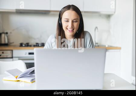 Smiling female student wearing wireless headset sits at desk in the kitchen home, watching online webinars on the laptop and taking notes, cheerful young woman working or studying on the distance Stock Photo