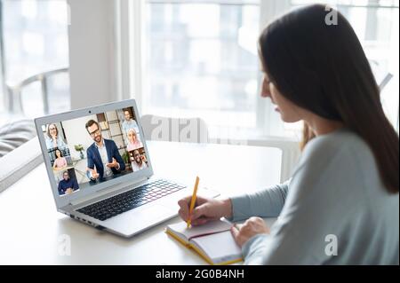 Young woman using laptop computer for watching online courses or webinars, female student takes a part in video conference, takes notes, writes down, e-learning and studying on the distance Stock Photo