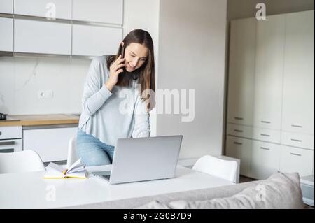 Smiling young woman using laptop computer for remote work from home, talks on the smartphone stands near desk and looks at the laptop screen, female freelancer has pleasant phone conversation Stock Photo