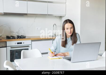 Executive freelance woman using laptop for remote work from home. Female student in casual wear sits at the desk in the kitchen, watching online webinars or training and taking notes, writes down Stock Photo