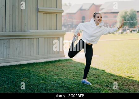 Pleased active young woman in hoodie and leggings prepares for running stratches and warms legs focused happily into distance listens music via