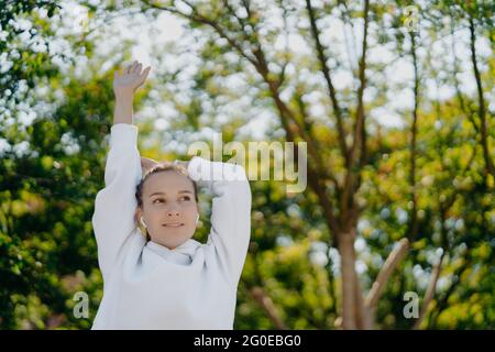 Relaxed beautiful young woman does stretching exercises outdoors keeps arms raised up focused into distance wears white hoodie listens music in