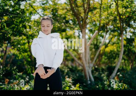 Horizontal shot of sporty European woman stretches hands and warms up before workout wears white sweatshirt andleggings poses against trees background Stock Photo