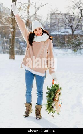 Happy female in outerwear carrying decorated Christmas wreath and standing on snow on sunny winter day Stock Photo