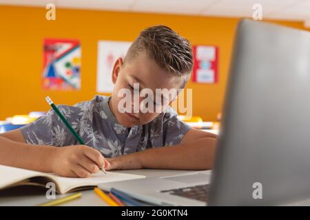 Caucasian boy sitting at a desk in classroom writing and using laptop Stock Photo