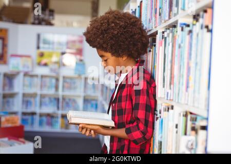 Happy african american schoolboy reading book standing in school library Stock Photo
